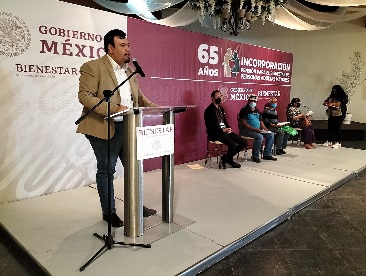Un hombre habla en un podio de la Secretaría de Bienestar ante una pancarta de incorporación a la Pensión para el Bienestar de Personas Adultas Mayores, con personas con cubrebocas sentadas al fondo