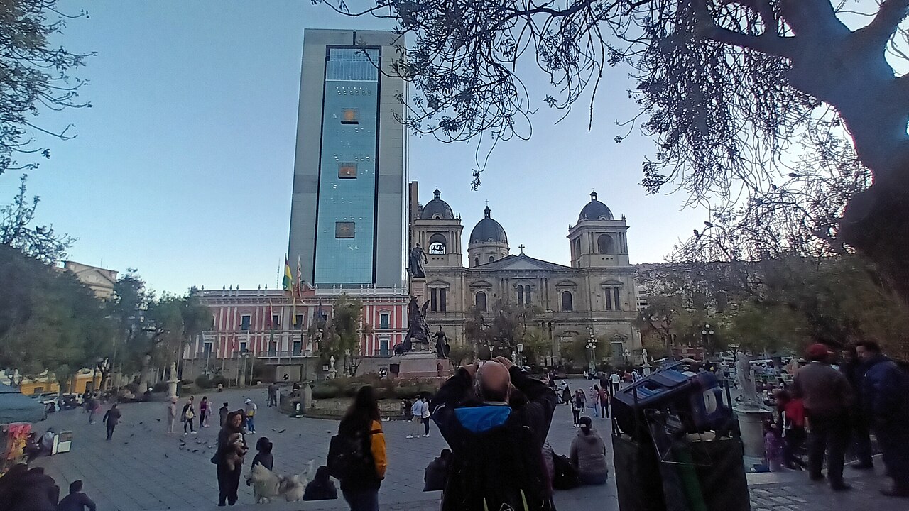 La plaza Murillo de La Paz, con la catedral, una torre de cristal y otros edificios al fondo y gente caminando por la plaza