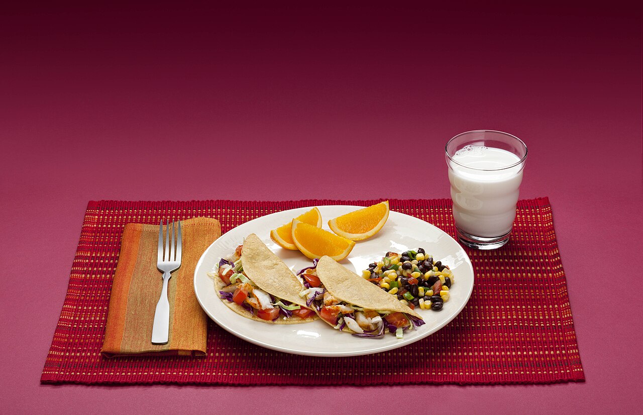 Un plato con una comida equilibrada: tacos, naranja, frijoles con maíz y un vaso de leche