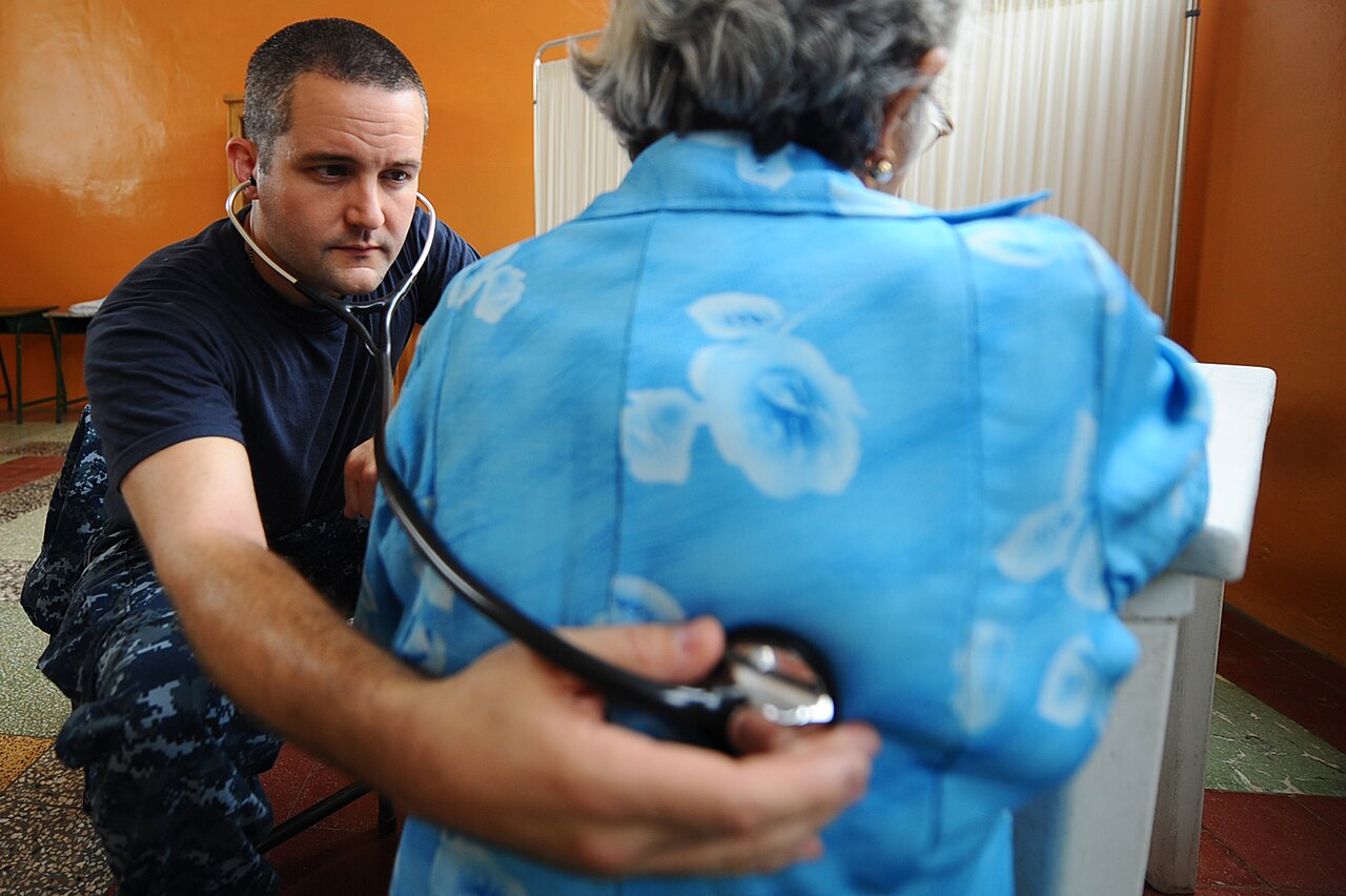 Un médico escucha con un estetoscopio la espalda de una paciente sentada y de espaldas, en una consulta