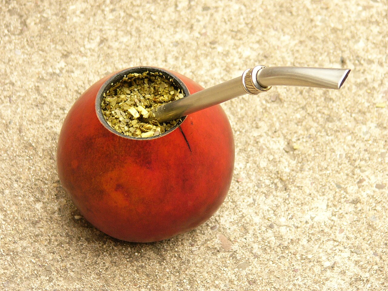 Calabaza roja llena de yerba mate con una bombilla metálica asomando, sobre un suelo de cemento