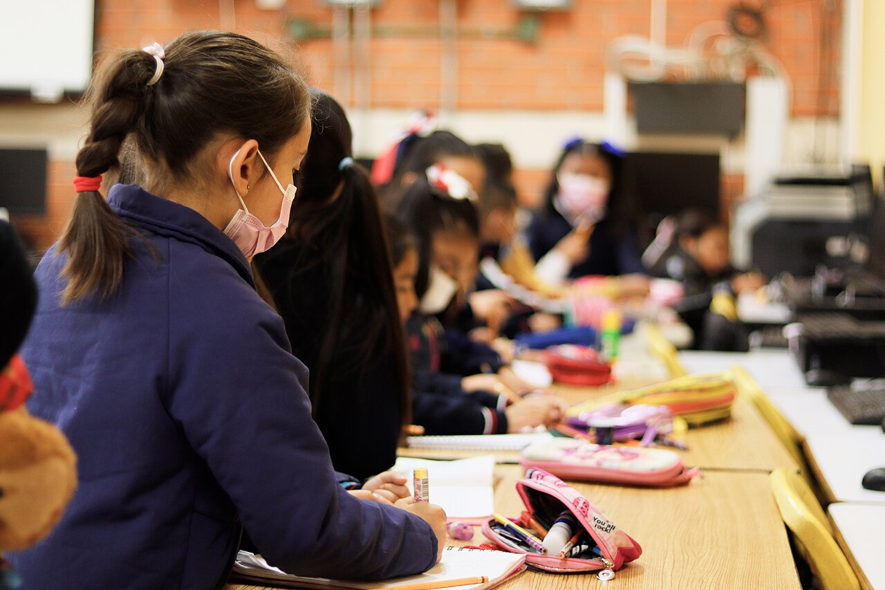 Niñas con uniforme escolar azul escriben en sus pupitres, con estuches y cuadernos sobre la mesa, en un salón de una escuela pública de la Ciudad de México