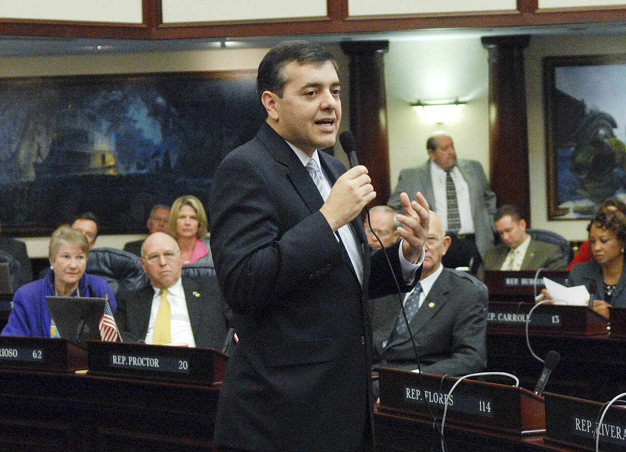 David Rivera habla con un micrófono en la mano en el pleno de la Cámara de Representantes de Florida; en el pupitre se lee «Rep. Rivera»