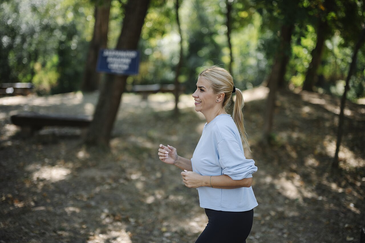 Una mujer sonríe mientras corre por un sendero de un parque arbolado, con un top azul claro y mallas oscuras
