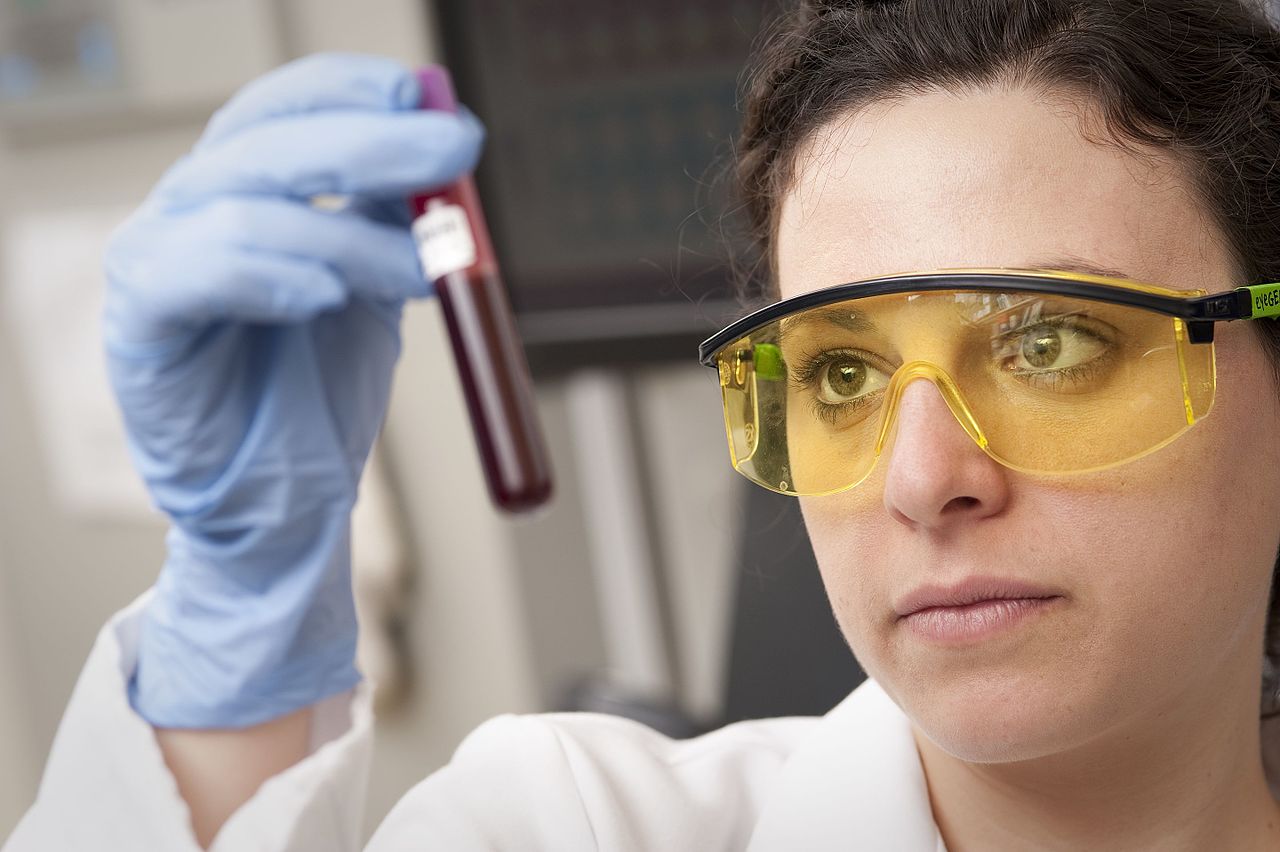 Una investigadora con bata, guantes y gafas de protección amarillas mira un tubo con una muestra de sangre que sostiene en alto