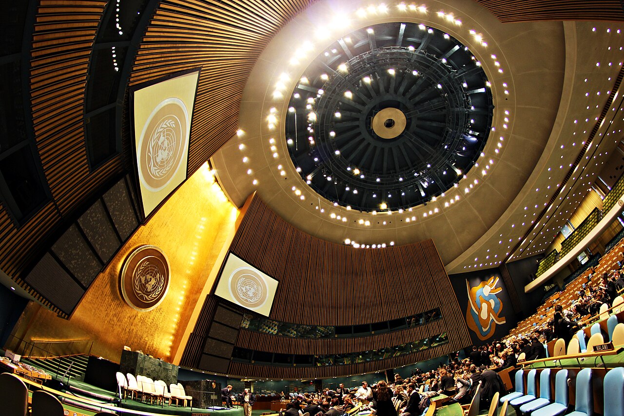 Interior del Salón de la Asamblea General de la ONU, en la sede de Nueva York, con el emblema de la organización sobre la tribuna y los asientos de las delegaciones