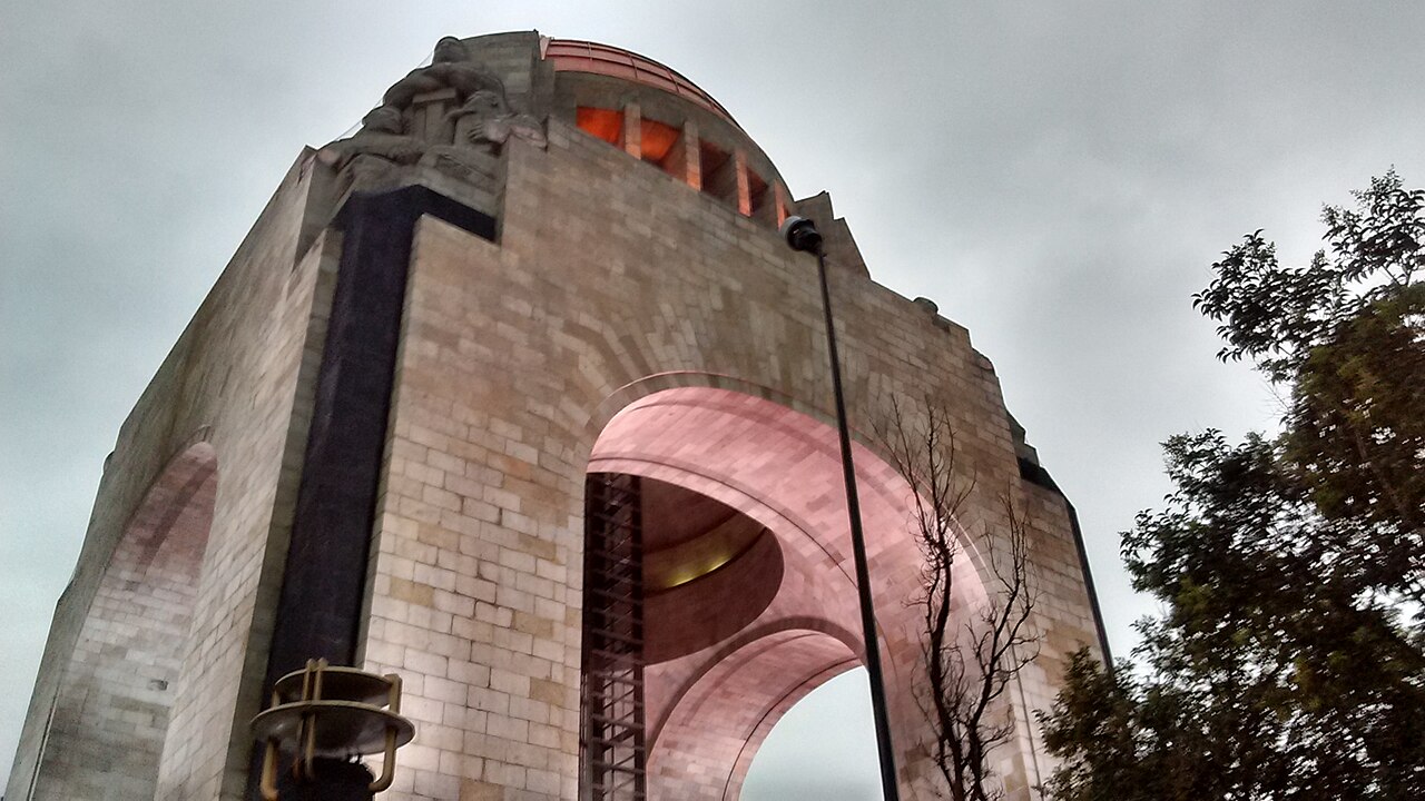 Vista desde abajo del Monumento a la Revolución en la Ciudad de México, con su arco de piedra y la cúpula iluminada bajo un cielo nublado