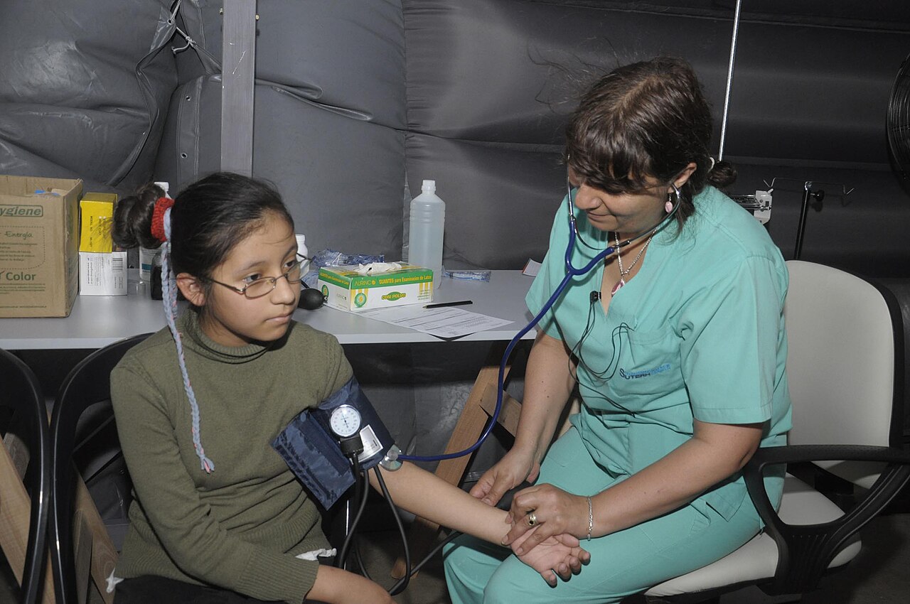 Una enfermera con fonendoscopio mide la tensión arterial a una niña sentada, con el manguito en el brazo, en una consulta improvisada