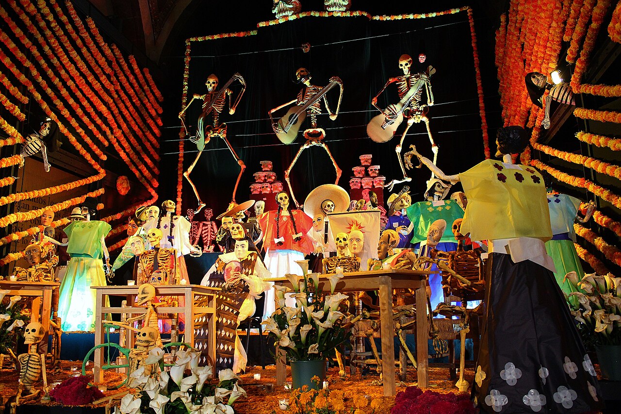 Altar monumental de Día de Muertos con esqueletos de cartón tocando guitarras, guirnaldas de flores de cempasúchil y mesas con catrinas
