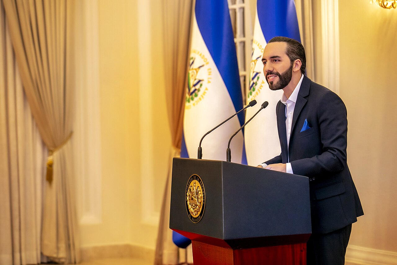 Nayib Bukele hablando ante dos micrófonos en un atril con el escudo de El Salvador, junto a dos banderas salvadoreñas