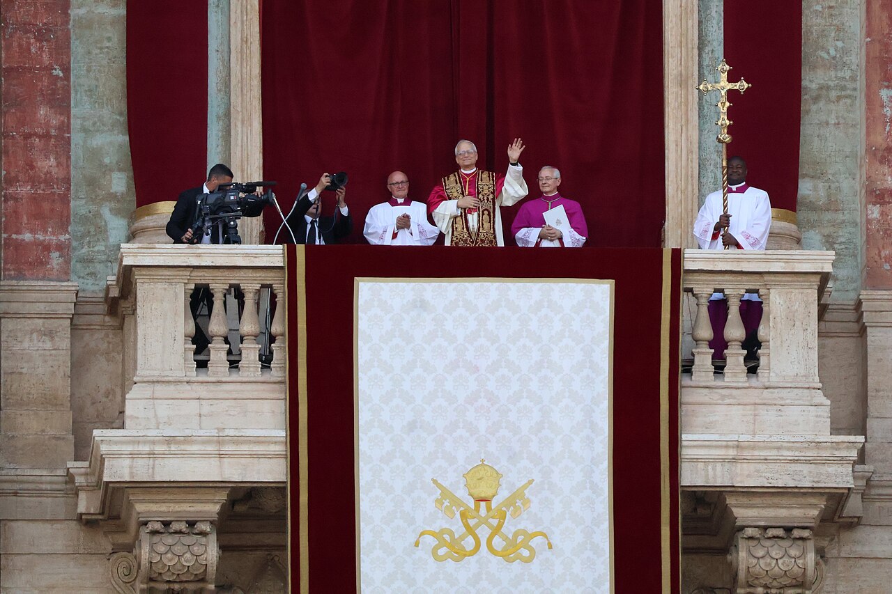 El papa León XIV saluda desde la logia de la basílica de San Pedro el 8 de mayo de 2025, acompañado por clérigos, con el escudo pontificio en el tapiz